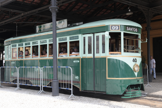 Bonde turístico verde, fechado, repleto de passageiros, em frente à Estação do Valongo no centro histórico de Santos