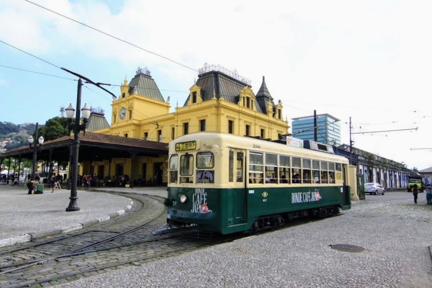 Bonde turístico japonês, bege e verde, fechado, repleto de passageiros, próximo à estação no centro histórico de Santos