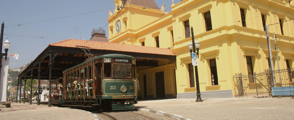 Bonde turístico aberto em frente à antiga estação ferroviária restaurada, de fachada amarela, no centro histórico de Santos