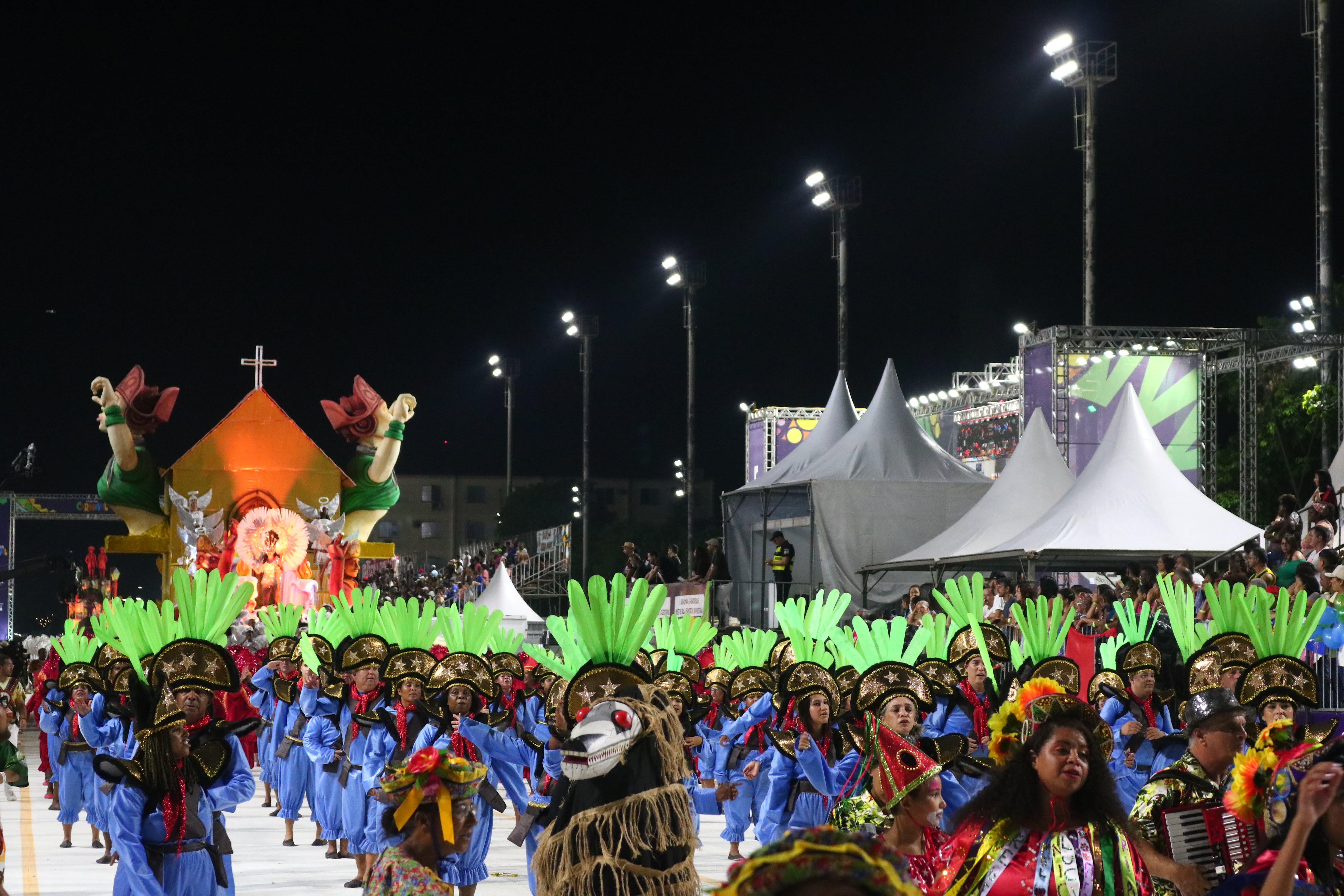 Desfile de carnaval à noite, com integrantes fantasiados em cores vibrantes, e um grande carro alegórico ao fundo