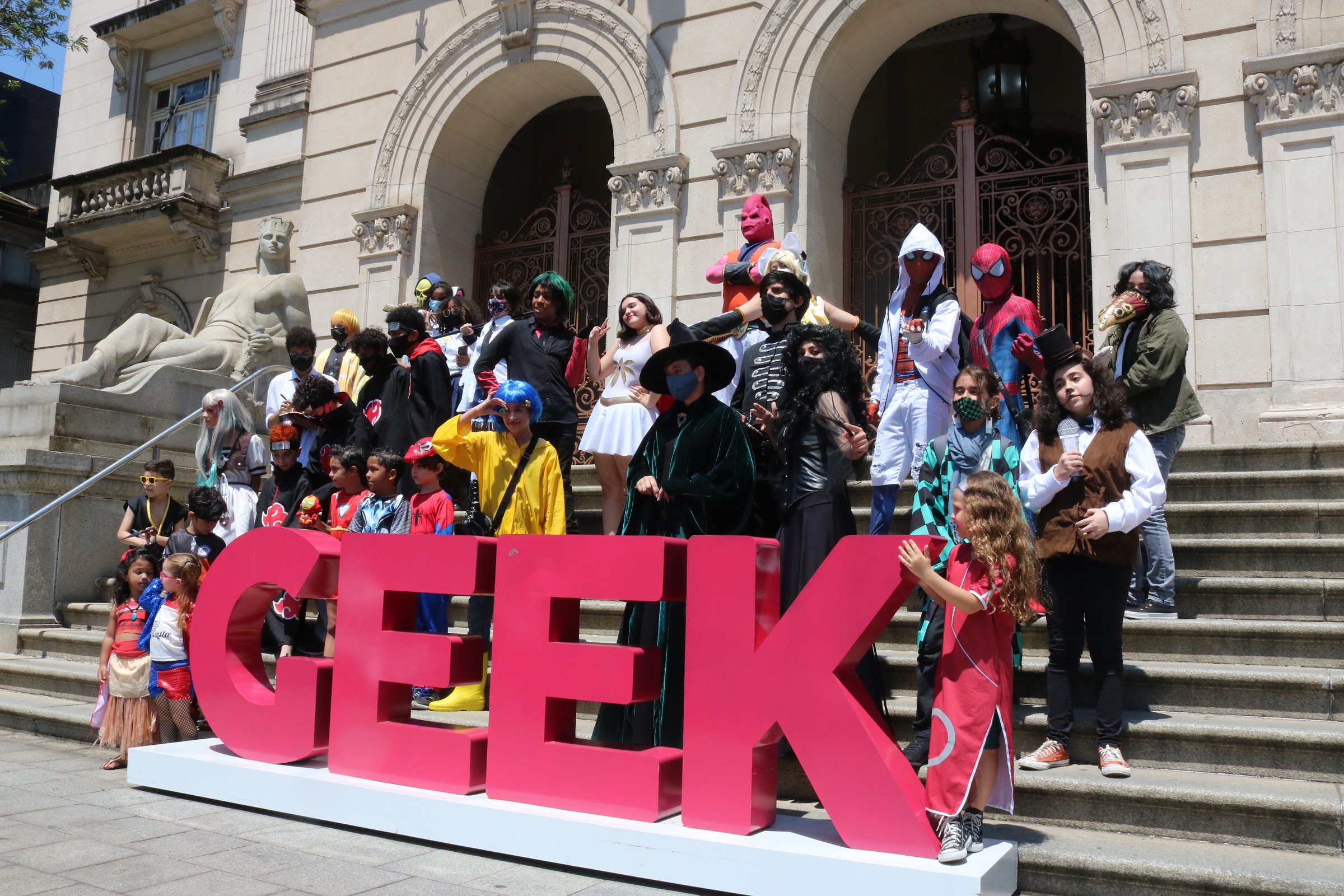 Diversos personagens de cosplay, junto a um letreiro Geek posicionado na escadaria do Paço Municipal de Santos