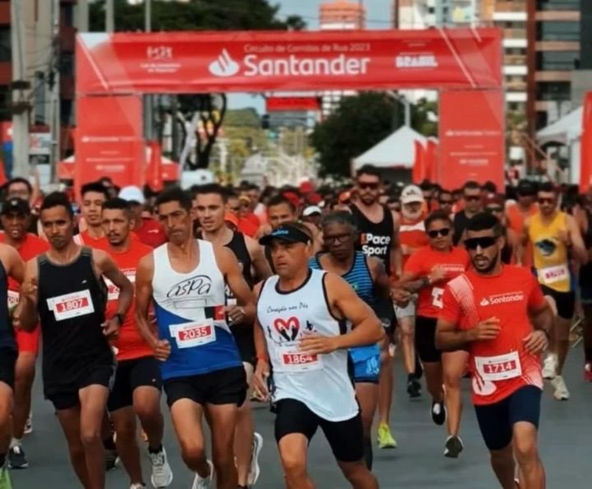 Grupo de corredores participando de uma prova de rua, com faixa de largada do evento esportivo ao fundo