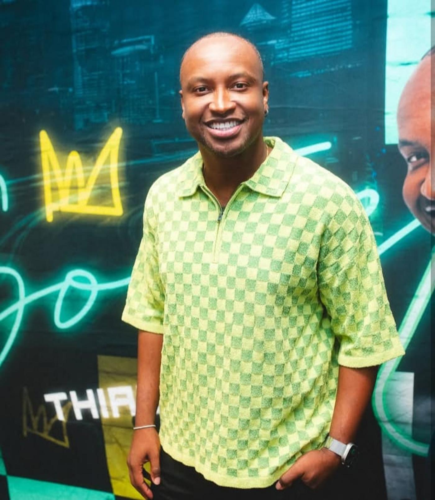 Cantor Thiaguinho, sorridente, com camiseta xadrez e fundo com letras neon