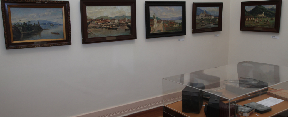 A imagem mostra o interior de uma sala expositiva, com paredes claras e iluminação discreta, típica de um espaço museológico. Na parede principal, observa-se uma sequência de pinturas emolduradas, dispostas de forma alinhada e equilibrada. As obras retratam paisagens e cenas urbanas, com destaque para áreas portuárias, igrejas, construções históricas e ambientes naturais, sugerindo registros artísticos de diferentes épocas ou vistas de uma mesma região.