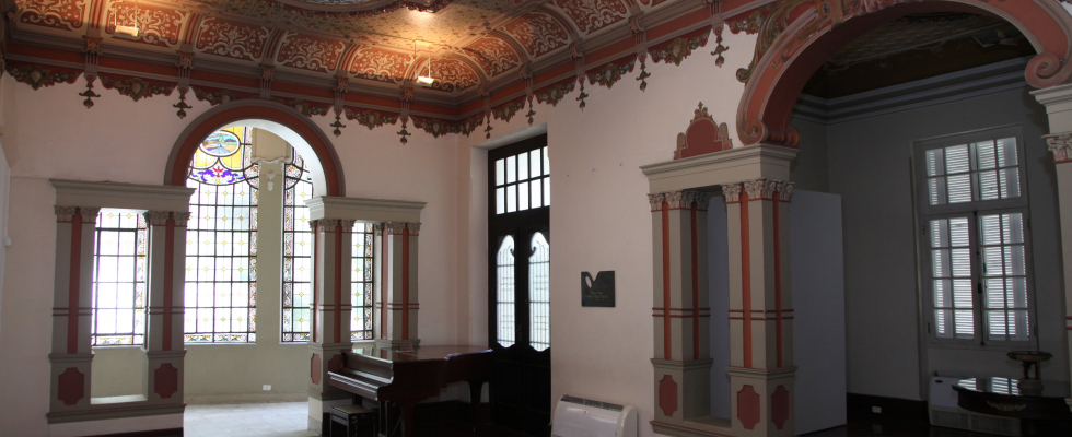 A imagem mostra um ambiente interno de um edifício histórico, marcado por rica ornamentação e atmosfera elegante. O espaço possui pé-direito alto e é valorizado por um teto decorado, com pinturas e relevos em tons quentes, além de pequenos detalhes pendentes que reforçam o caráter artístico do conjunto.