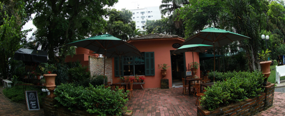 A imagem retrata um espaço acolhedor ao ar livre, integrado a um jardim arborizado, onde funciona um bistrô instalado em um anexo histórico. A construção de tons terrosos e rosados, com portas e janelas em madeira pintadas de verde, remete a uma arquitetura charmosa e intimista, em harmonia com o entorno natural.