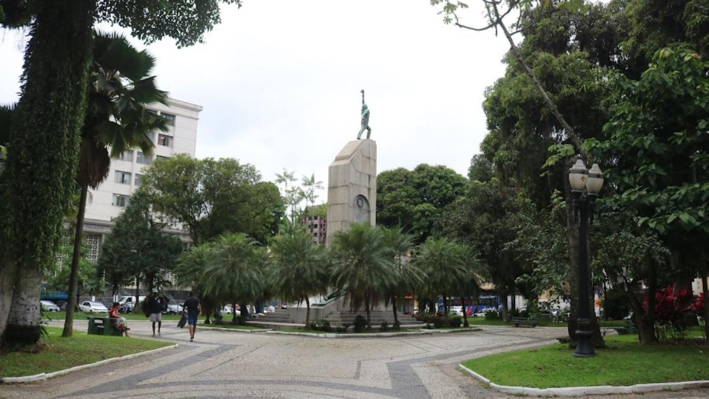 Praça arborizada com monumento central, rodeada por árvores, jardins e caminhos de pedra
