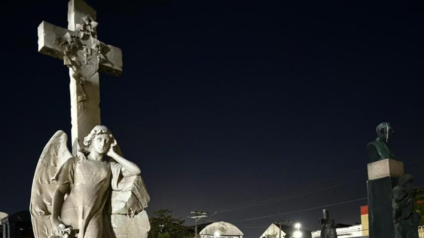 A imagem mostra uma cena noturna no Cemitério do Paquetá, em Santos, registrada durante um passeio guiado. Em primeiro plano, destaca-se uma imponente escultura de anjo em mármore claro, em posição contemplativa ao lado de uma grande cruz ornamentada. O anjo segura flores, símbolo de luto e memória, enquanto sua expressão serena transmite paz. O túmulo onde está a escultura é robusto, em pedra clara, com inscrições na lápide.