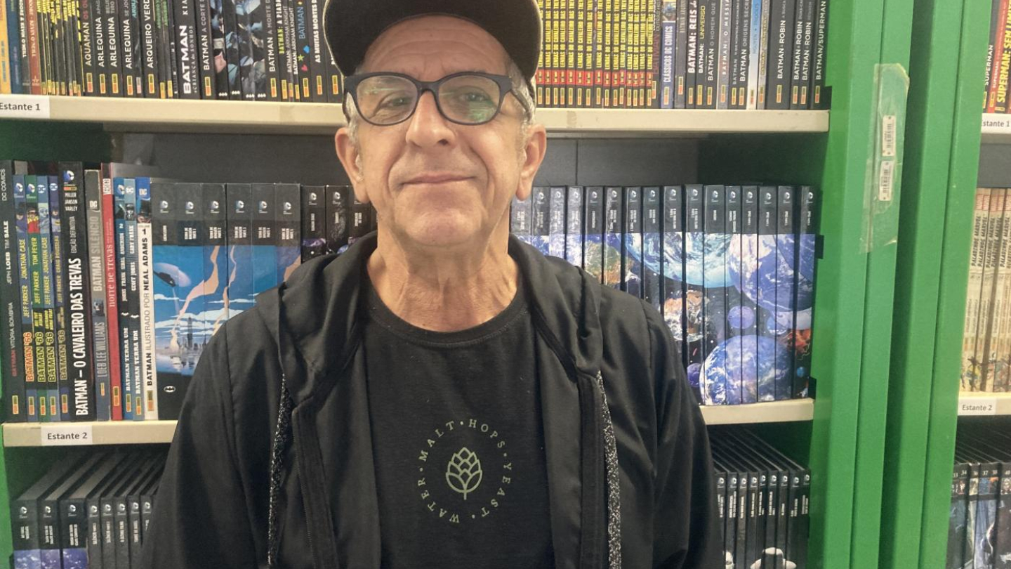 Foto do ilustrador Osvaldo Da Costa, com diversos livros ao fundo