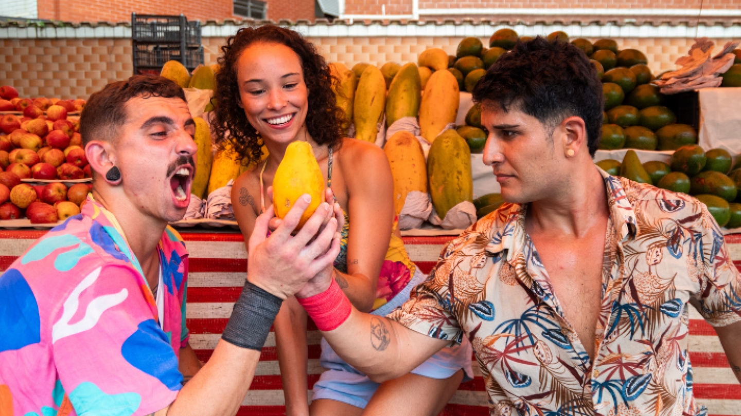 Três pessoas interagem de forma divertida em uma barraca de frutas, com papaias e mangas ao fundo, criando uma cena vibrante e espontânea de mercado