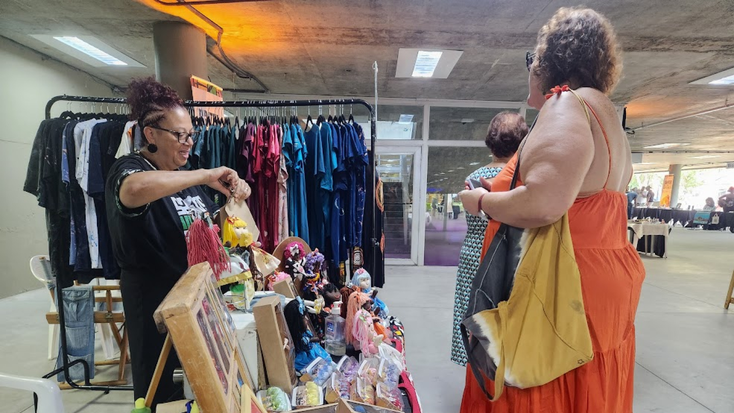 Uma vendedora em uma feira de artesanato em um espaço interno, mostrando bonecas e produtos artesanais para uma cliente vestindo um vestido laranja