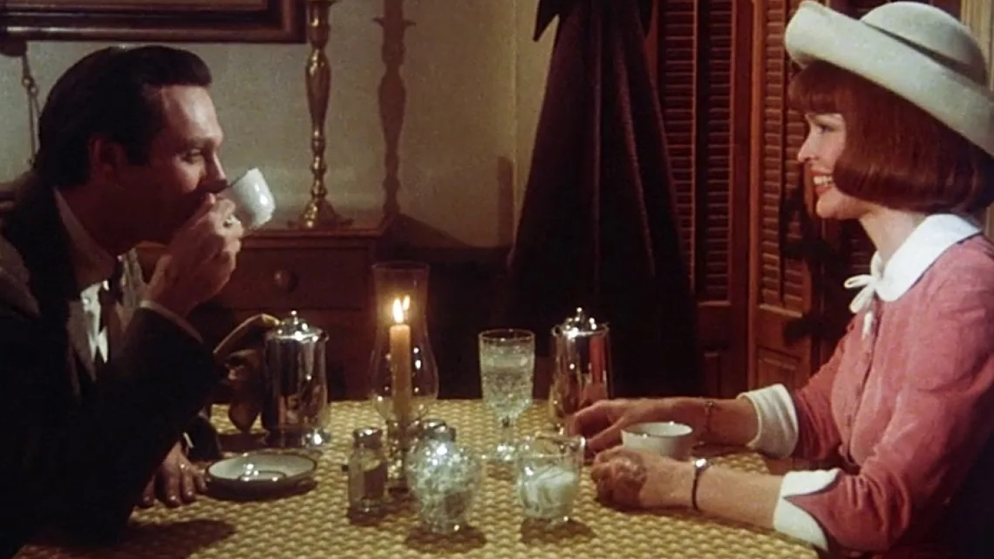 Casal, com roupas de época, toma bebida à luz de velas, num ambiente intimista