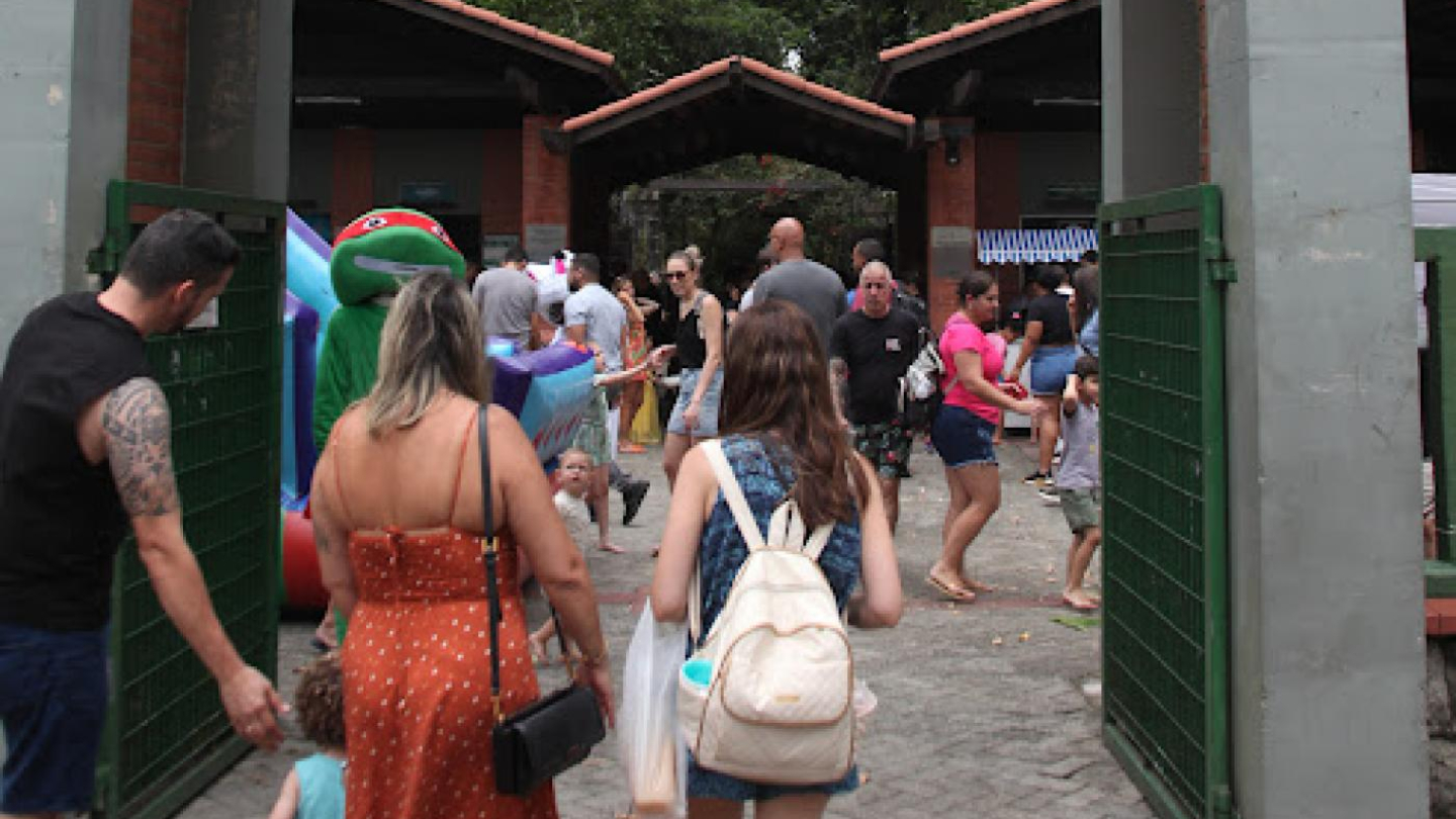A foto mostra a entrada do Orquidário de Santos, identificada por um grande pórtico com a placa “Parque Zoobotânico – Orquidário Santos”, acompanhada de ícones de animais, plantas e pessoas. Os portões verdes estão abertos, permitindo a passagem de diversos visitantes.