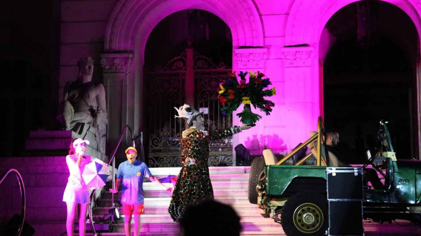 A imagem mostra uma apresentação artística realizada em frente a um prédio histórico de arquitetura clássica, iluminado com luzes rosa e roxas que destacam seus arcos e esculturas.  No centro da cena, um artista usando um figurino exuberante — uma roupa longa e brilhante, decorada com luzes e detalhes reluzentes, além de um chapéu adornado com penas — segura um grande buquê de flores coloridas, levantado de forma teatral.