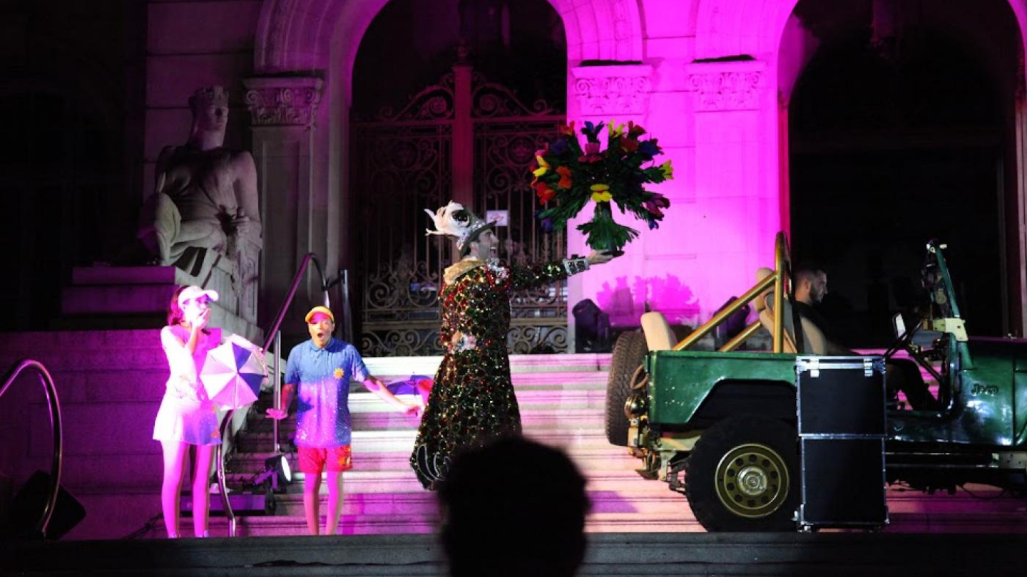 A imagem mostra uma apresentação artística realizada em frente a um prédio histórico de arquitetura clássica, iluminado com luzes rosa e roxas que destacam seus arcos e esculturas.  No centro da cena, um artista usando um figurino exuberante — uma roupa longa e brilhante, decorada com luzes e detalhes reluzentes, além de um chapéu adornado com penas — segura um grande buquê de flores coloridas, levantado de forma teatral.