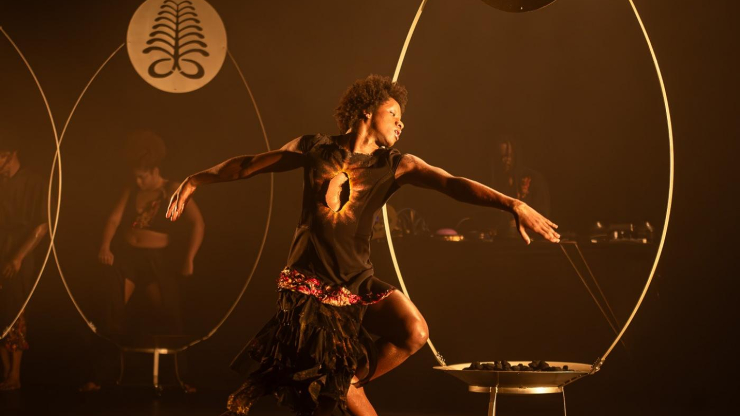 A imagem mostra uma cena de espetáculo de dança contemporânea, marcada por iluminação quente em tons de dourado e âmbar, criando um clima intenso e quase ritualístico.