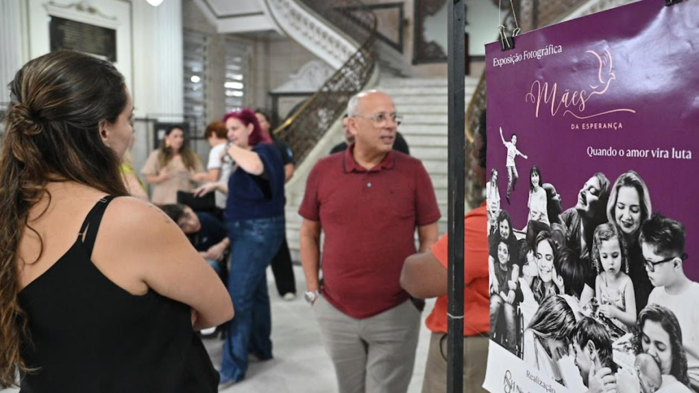 A imagem mostra o interior de um prédio histórico — com colunas brancas, piso claro e uma grande escadaria ao fundo — durante a abertura de uma exposição fotográfica. No primeiro plano, à direita, aparece um banner roxo da mostra “Mães da Esperança”, com o subtítulo “Quando o amor vira luta”. O cartaz exibe diversas fotografias em preto e branco de mães e filhos em momentos de carinho e superação.