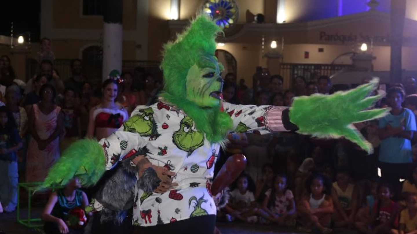 A imagem mostra uma apresentação artística ao ar livre, realizada à noite, com um performer vestido como o Grinch, personagem fictício conhecido por sua aparência verde e comportamento travesso.