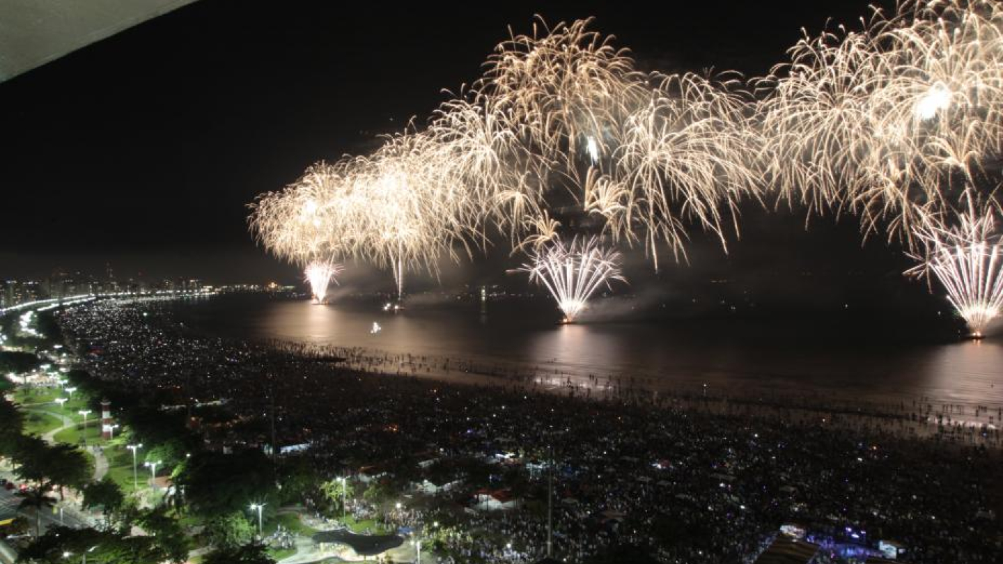 Vista aérea noturna da orla de Santos tomada por uma multidão, enquanto fogos de artifício dourados iluminam o céu e se refletem no mar, criando um cenário festivo e vibrante.