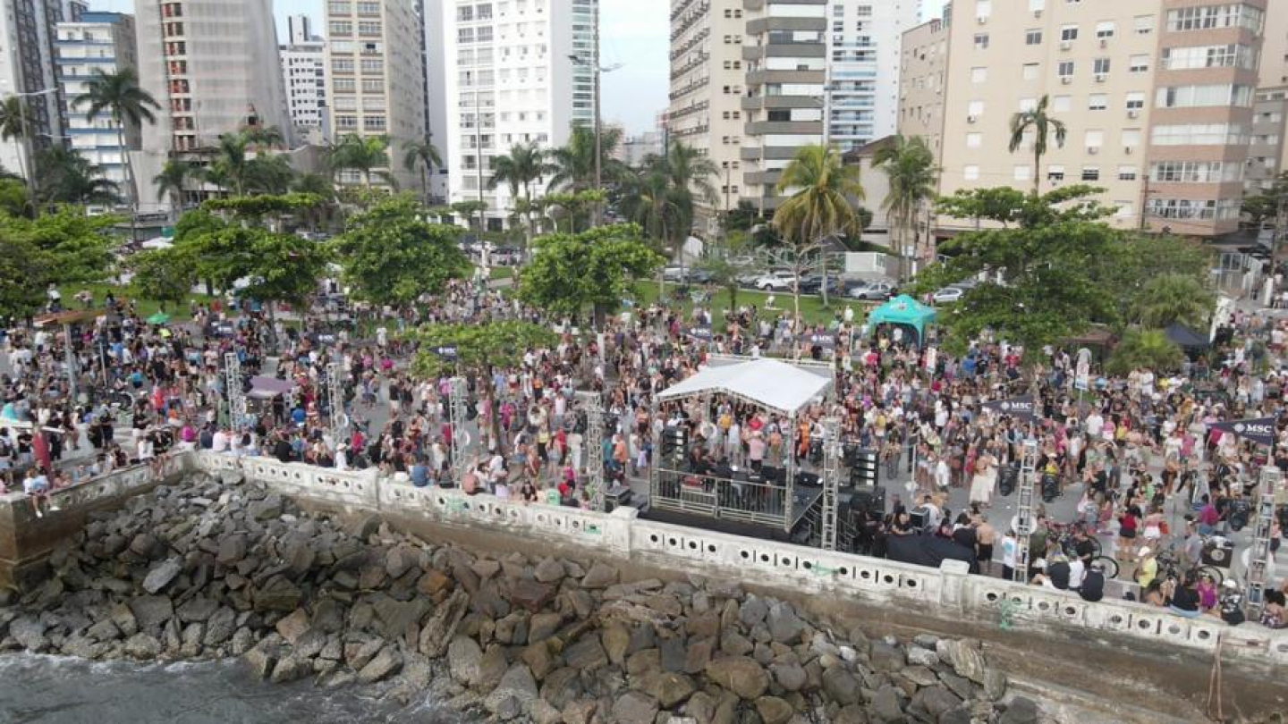 A imagem mostra uma grande concentração de pessoas reunidas em um evento ao ar livre na orla de Santos. A foto é aérea e destaca uma multidão ocupando toda a área do calçadão, próximo ao mar. No canto inferior, aparecem as pedras do paredão costeiro, onde as ondas batem suavemente.