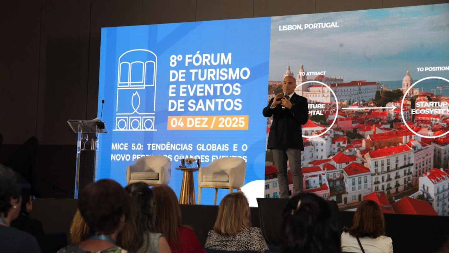 A imagem mostra uma palestra durante o 8º Fórum de Turismo e Eventos de Santos, realizado em 4 de dezembro de 2025. No palco, há um grande painel dividido em duas partes: à esquerda, a identidade visual oficial do evento, em azul, com o título e o tema “MICE 5.0: Tendências globais e o novo perfil dos grandes eventos”; à direita, uma foto aérea de Lisboa, Portugal, destacando elementos como “Future Capital” e “Startup Ecosystem”.