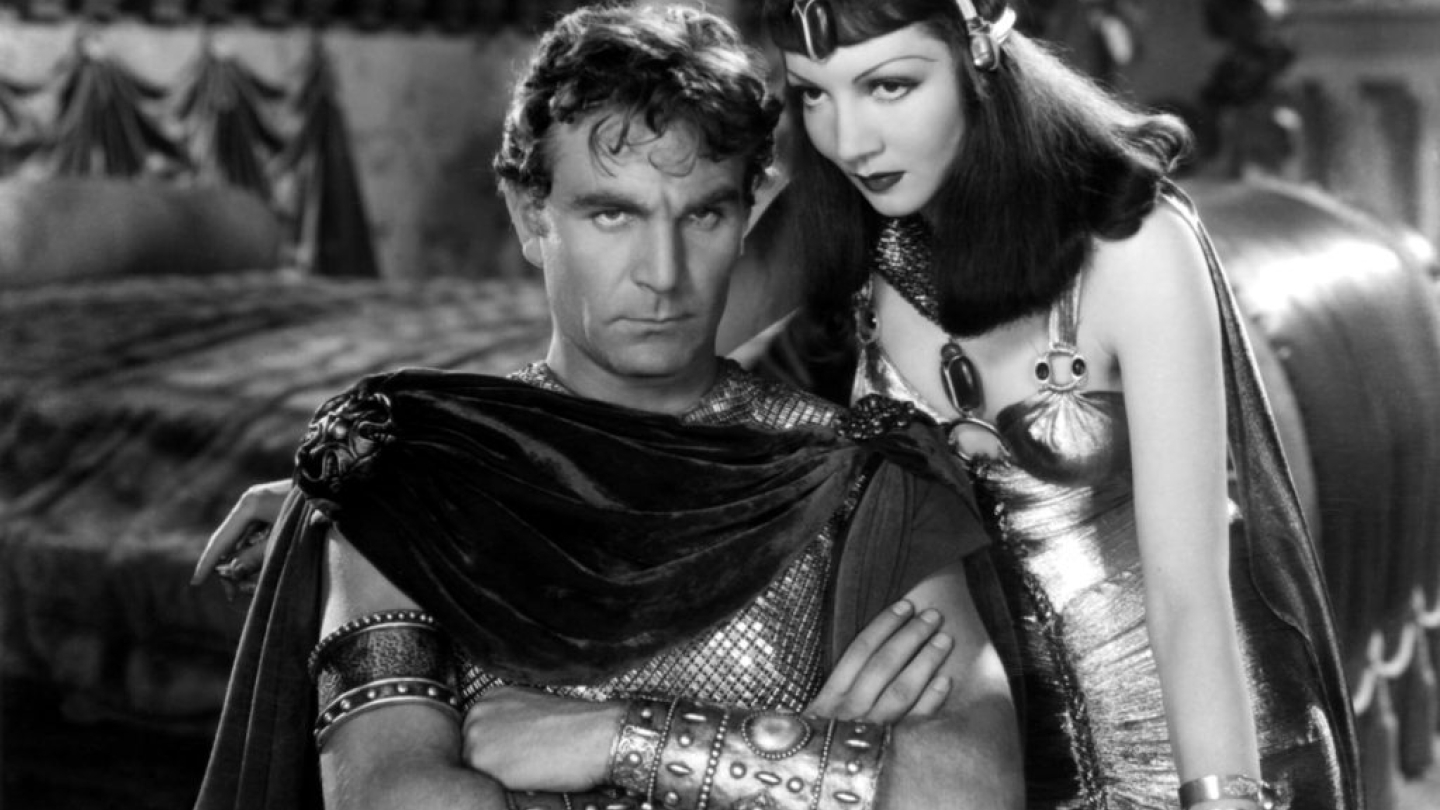 Fotografia em preto e branco do filme Cleópatra, mostrando a rainha do Egito com trajes luxuosos inclinada sobre um homem com armadura romana e braçadeiras ornamentadas