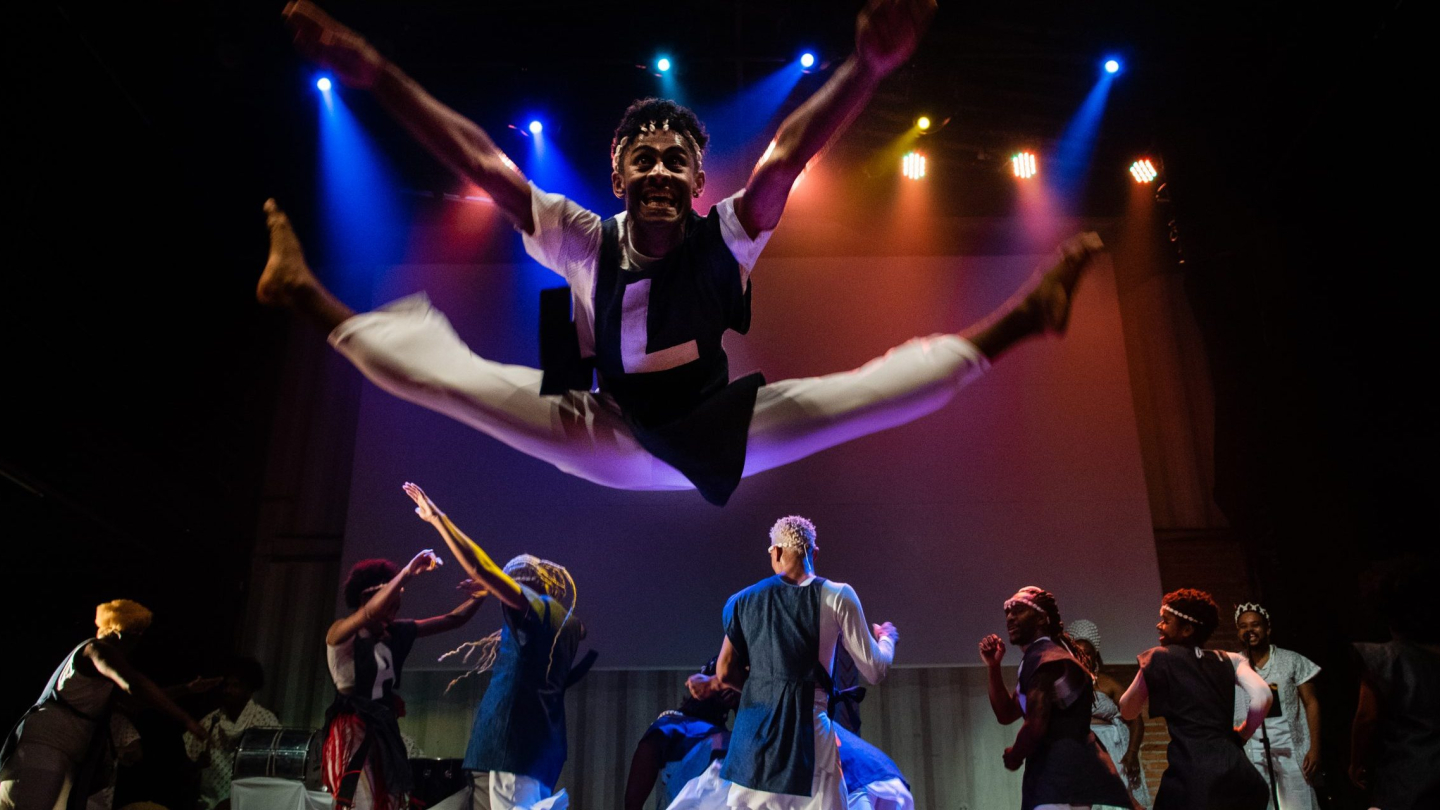 Um bailarino executa um salto acrobático com as pernas abertas no centro do palco, enquanto outros artistas dançam ao fundo sob luzes coloridas de um espetáculo teatral