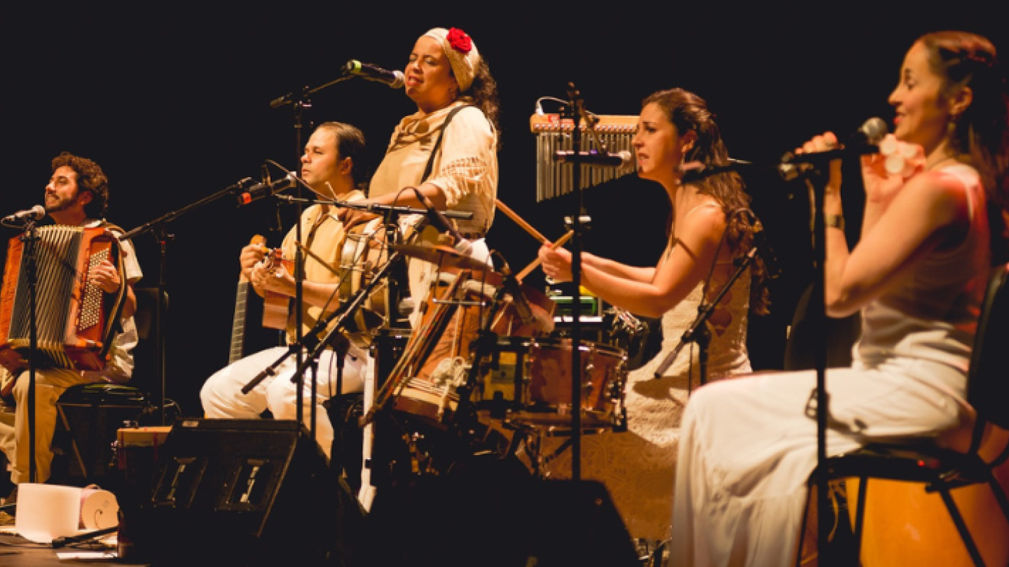 Um grupo de cinco músicos se apresenta no palco de um teatro, utilizando instrumentos como sanfona, cavaquinho e percussão