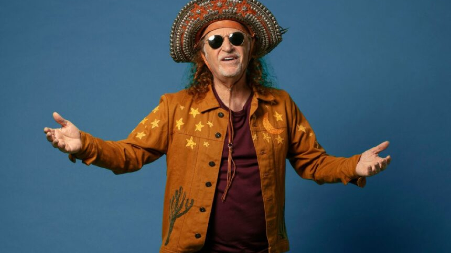 O cantor brasileiro Alceu Valença aparece sorridente com os braços abertos, vestindo uma jaqueta marrom decorada com estrelas e um tradicional chapéu de couro de cangaceiro contra um fundo azul