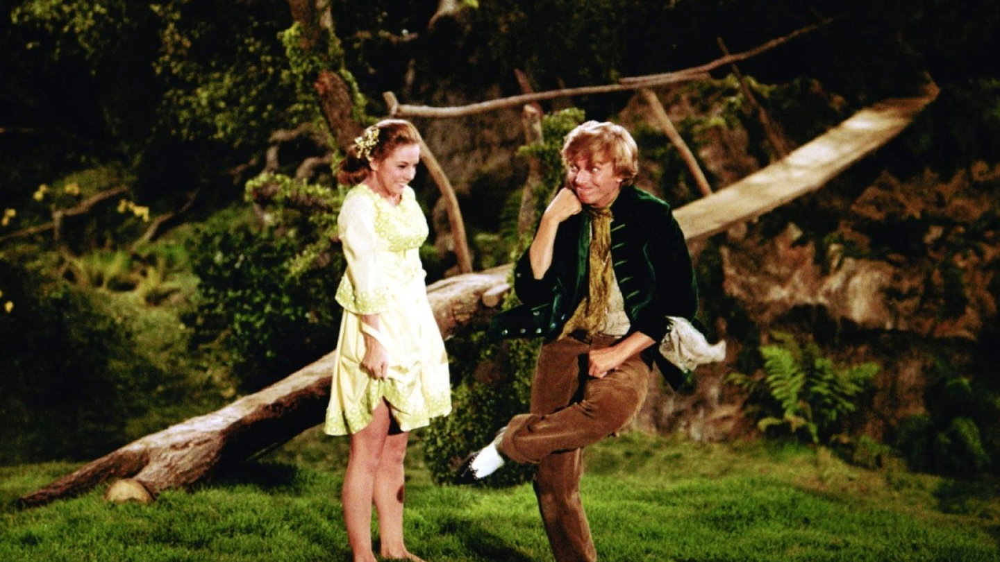 Uma mulher em um vestido claro observa um homem com traje de época dançar de forma descontraída em um cenário natural arborizado