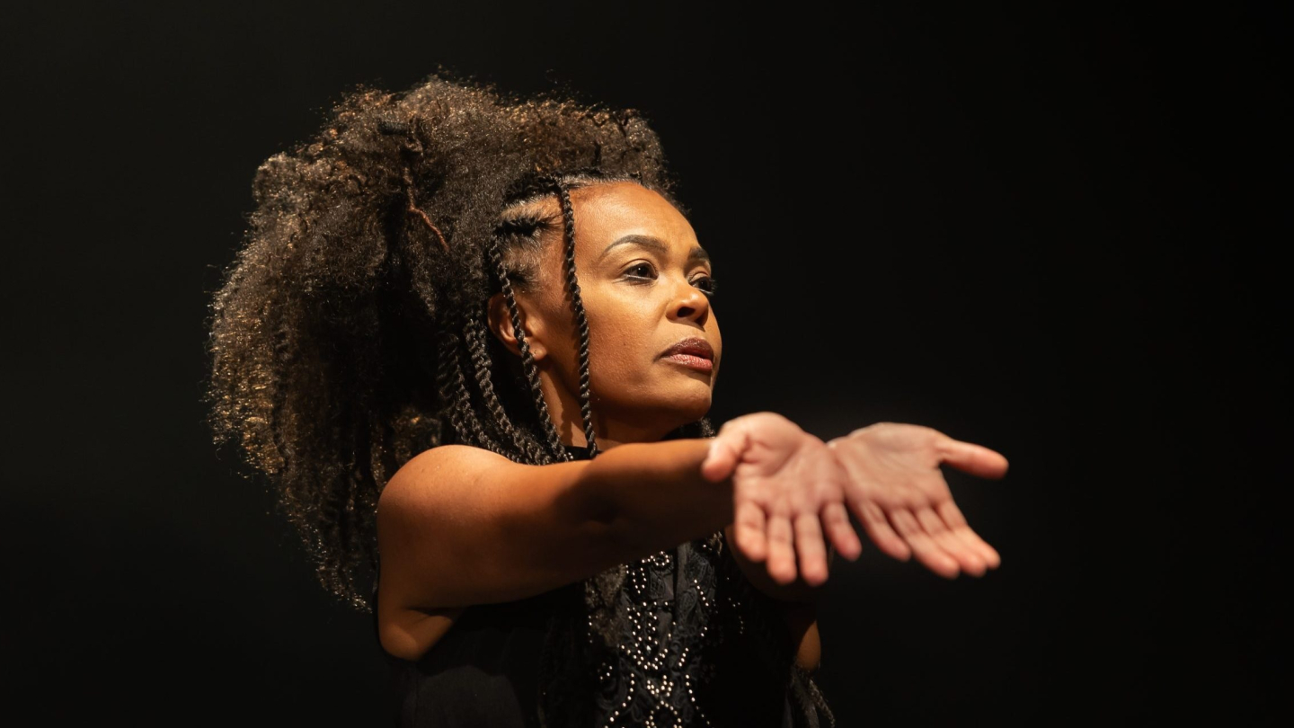 Uma mulher negra com um penteado volumoso e tranças laterais estende os braços para a frente com as palmas das mãos voltadas para cima em uma expressão performática sobre um fundo escuro