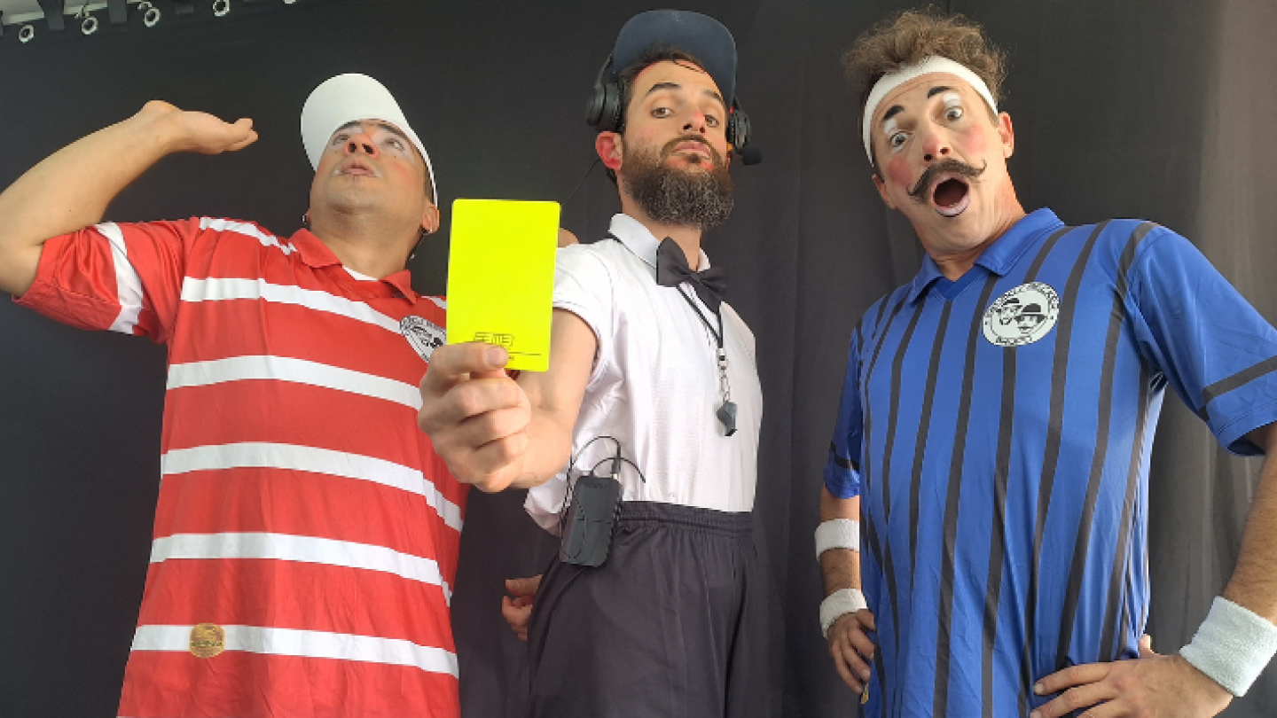 Três artistas caracterizados como personagens cômicos de esportes posam para a foto, com o homem ao centro segurando um cartão amarelo em destaque