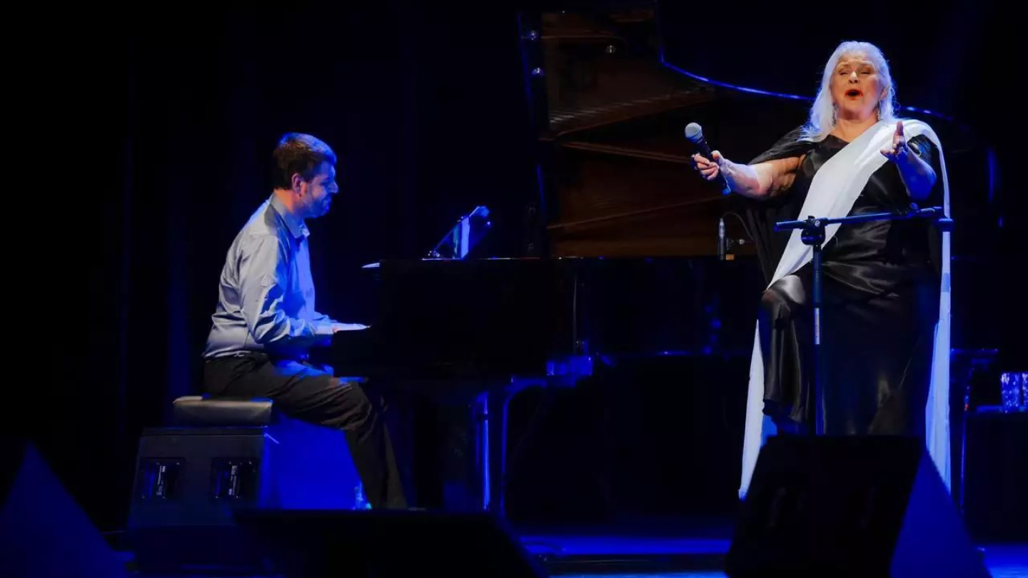 A cantora Fafá de Belém apresenta-se intensamente no palco sob luz azul, acompanhada por um pianista ao piano de cauda