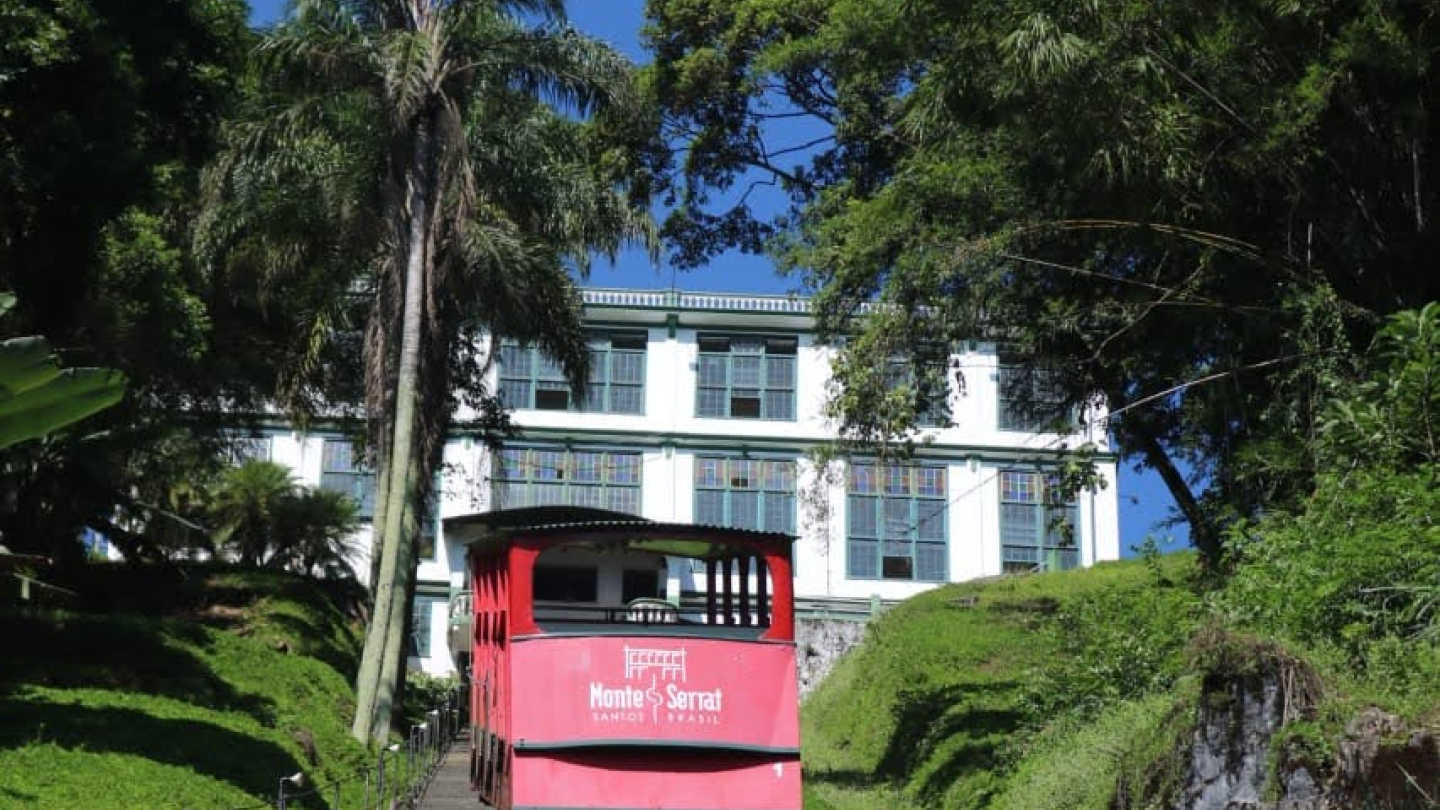 A imagem retrata o icônico bonde vermelho do Monte Serrat, em Santos, subindo os trilhos do funicular em meio à vegetação, com o histórico edifício do antigo cassino ao fundo