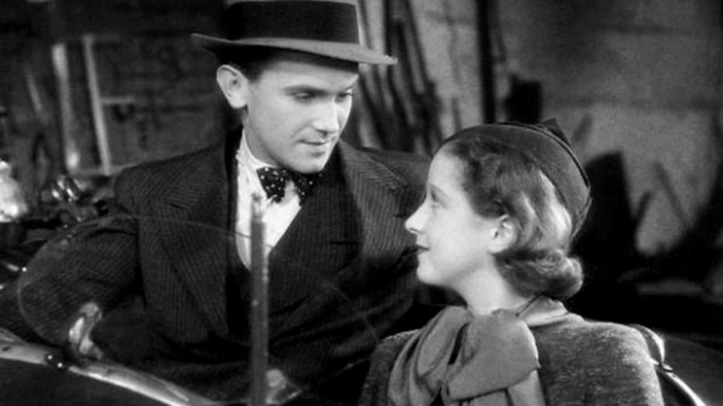 Uma cena em preto e branco mostra um homem de chapéu e gravata borboleta olhando para uma mulher em um ambiente que sugere o interior de um carro antigo