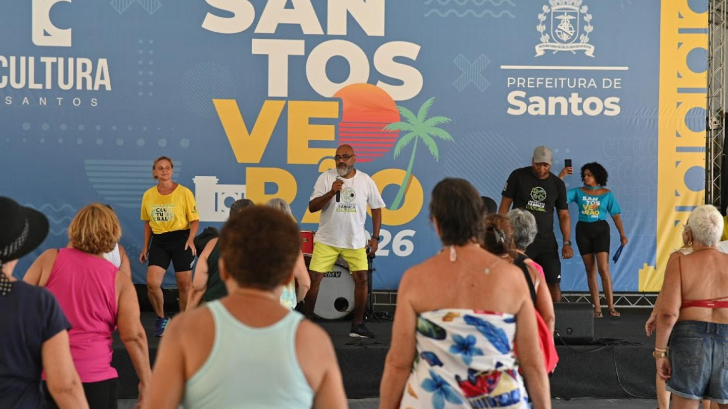 Um instrutor de ginástica lidera uma aula de dança para um grupo de pessoas em um palco sob uma tenda do evento "Santos Verão 2026"