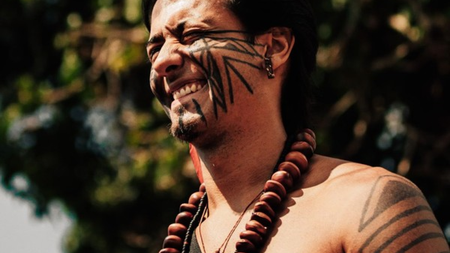 Retrato em close de um homem indígena sorrindo, exibindo pinturas faciais e corporais geométricas tradicionais e usando colares de sementes