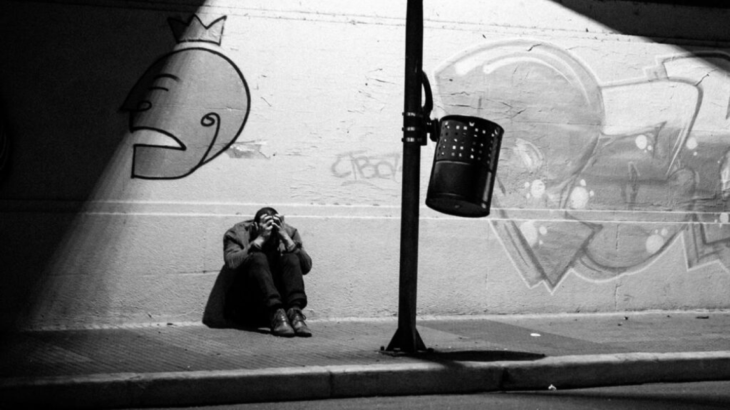 Uma pessoa aparece sentada no chão de uma calçada com as mãos na cabeça, em uma composição melancólica em preto e branco com grafites ao fundo