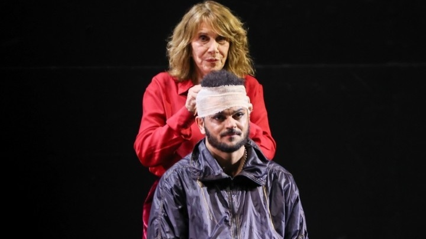 Uma mulher em uma camisa vermelha ajusta uma bandagem na cabeça de um homem sentado à sua frente, contra um fundo preto