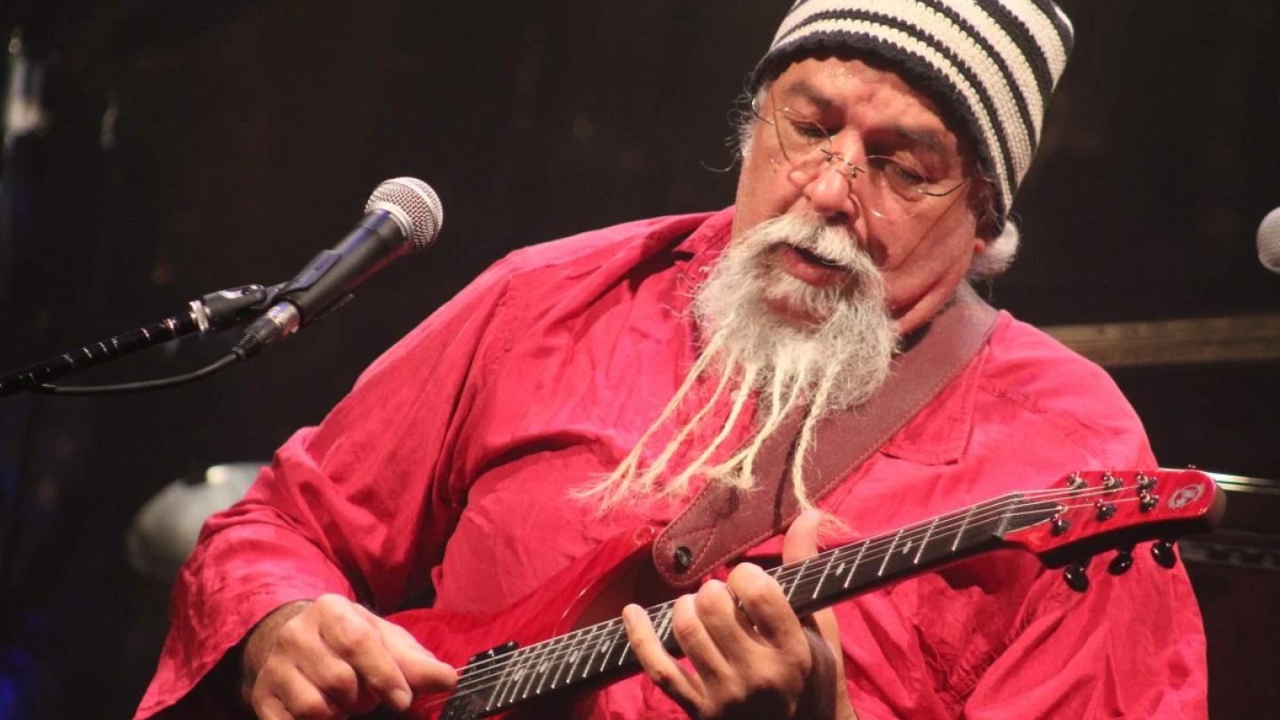 O multi-instrumentista Arismar do Espírito Santo aparece concentrado tocando uma guitarra vermelha, vestindo uma camisa da mesma cor e seu característico gorro listrado