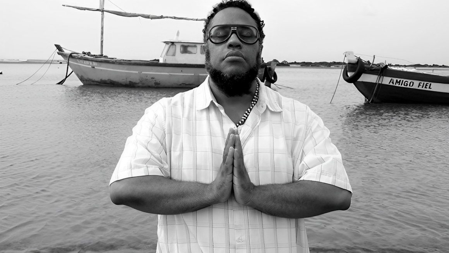 O músico BNegão aparece em uma fotografia em preto e branco, com as mãos postas em sinal de prece e olhos fechados, tendo barcos de pesca e o mar ao fundo