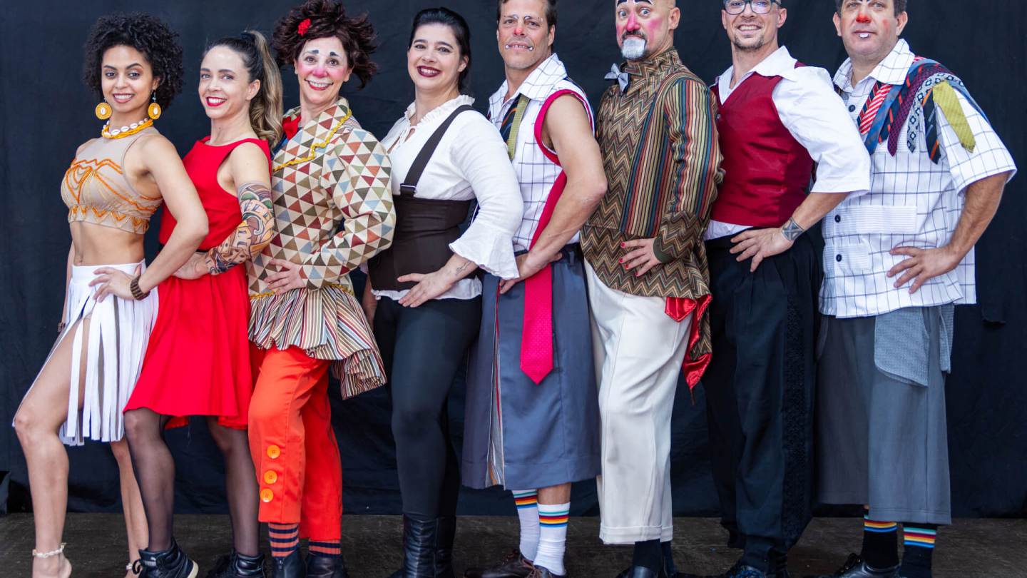 Um grupo de oito artistas circenses e performáticos posa sorridente em linha, exibindo figurinos coloridos, maquiagens de palhaço e trajes ecléticos de cabaré sobre um fundo escuro
