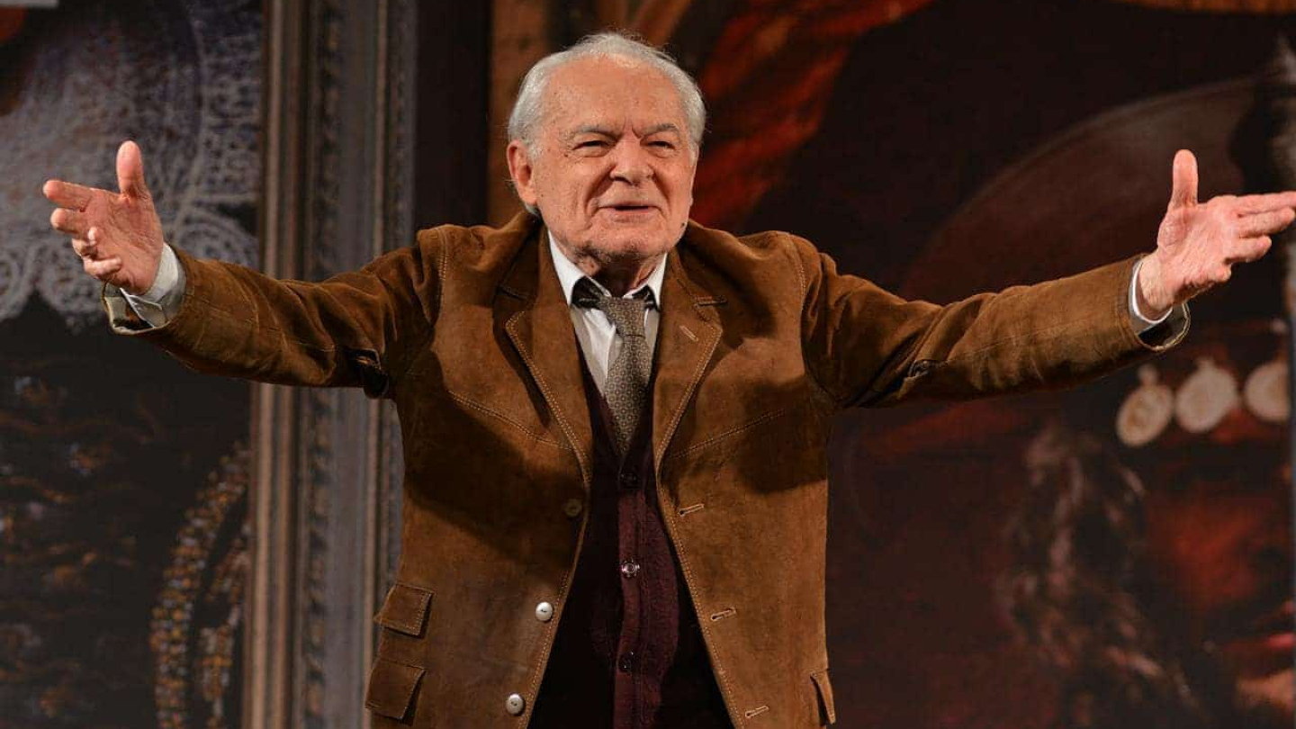O ator brasileiro Othon Bastos aparece de braços abertos e sorridente no palco, vestindo um casaco de camurça marrom sobre um colete e gravata