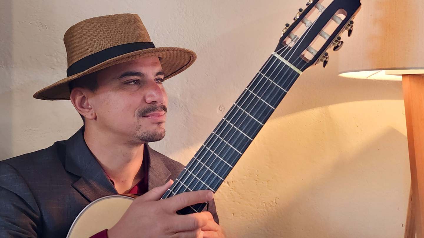 Um músico de chapéu e paletó segura um violão em um ambiente acolhedor e iluminado, transmitindo uma expressão serena e contemplativa