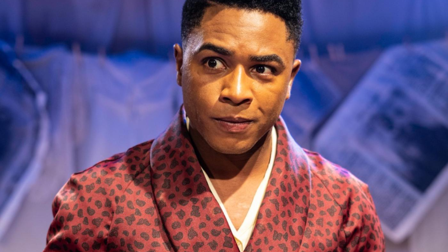 A imagem mostra o ator Sidney Santiago em cena, vestindo um roupão de seda bordô com estampa de leopardo e segurando três pequenos livros vermelhos amarrados com um laço preto