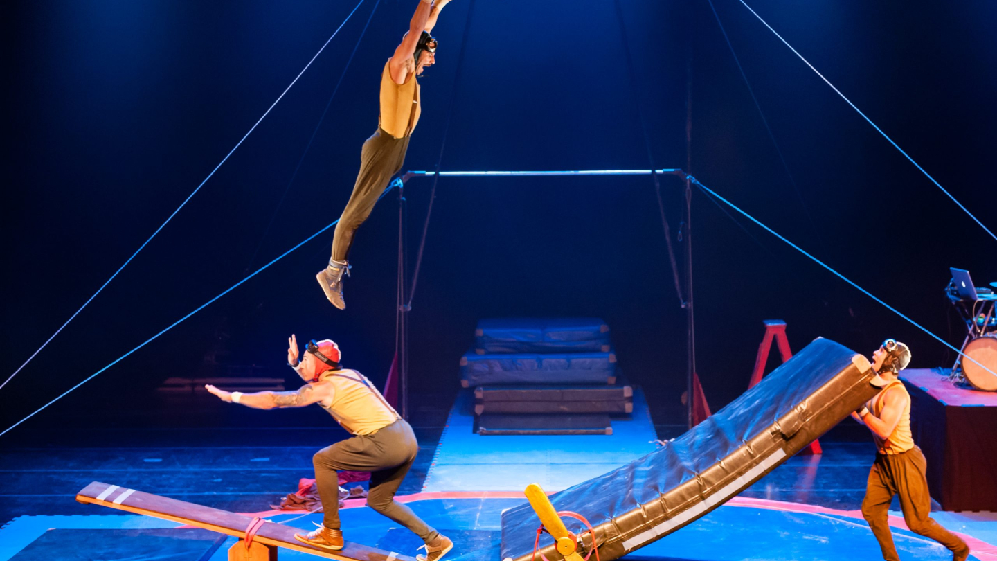 Acrobatas realizam uma performance circense dinâmica sob luzes azuis, com um artista saltando de uma gangorra enquanto outro posiciona um colchão de proteção no palco