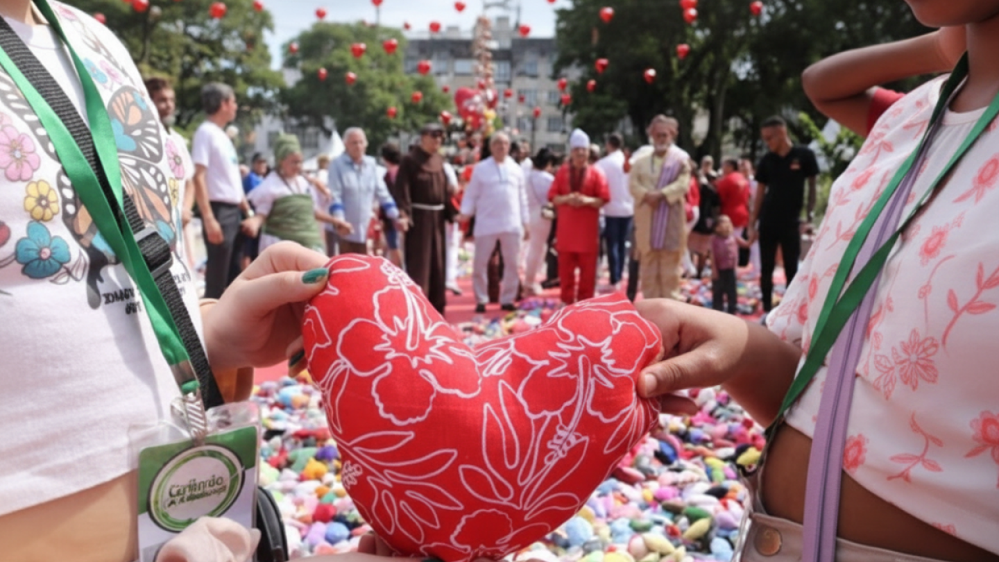 Duas jovens, com os rostos parcialmente ocultos, seguram juntas uma almofada em formato de coração vermelho com estampas florais brancas, diante de uma multidão reunida em uma praça decorada com balões vermelhos e um tapete de pequenos objetos coloridos