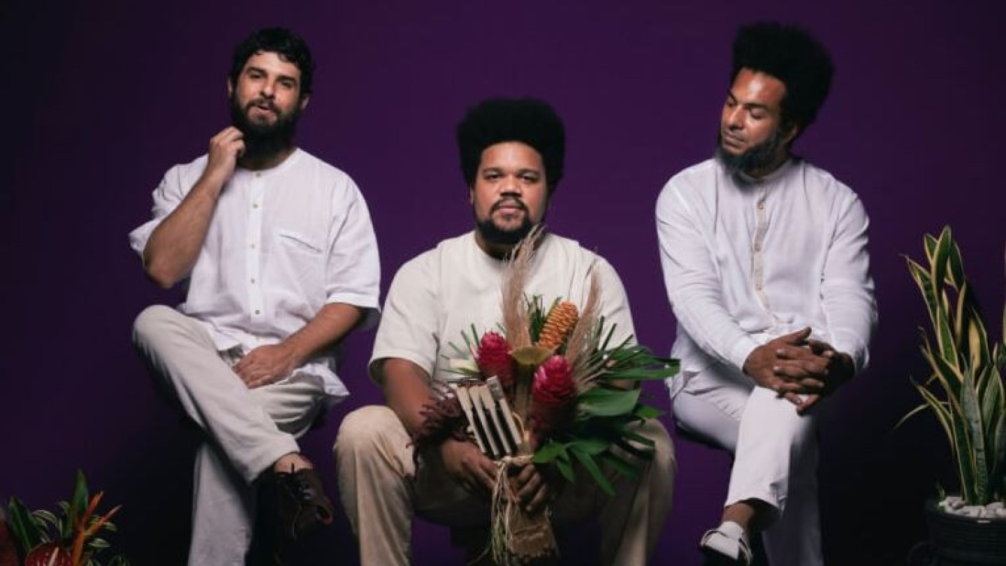 Os três integrantes do Amaro Freitas Trio posam sentados com roupas claras contra um fundo roxo vibrante, acompanhados por arranjos de plantas tropicais