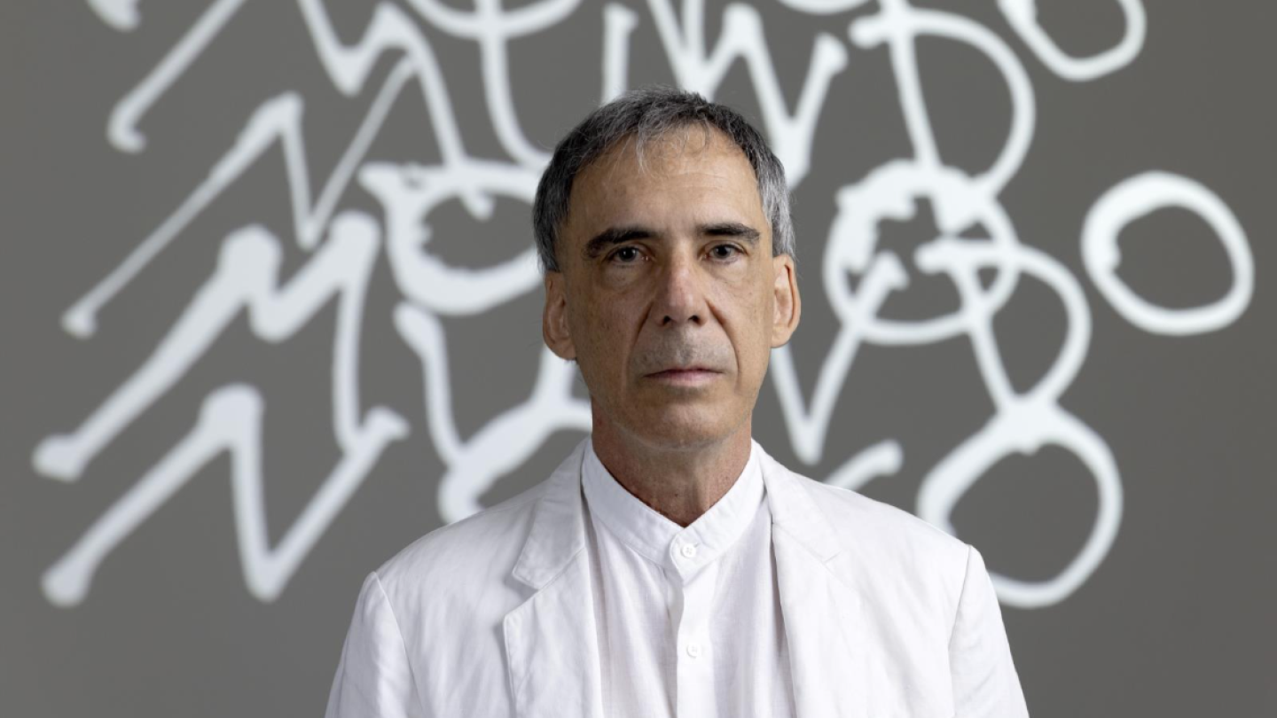 O músico e poeta Arnaldo Antunes posa de branco diante de um fundo cinza com grafismos brancos que formam a expressão "Novo Mundo"