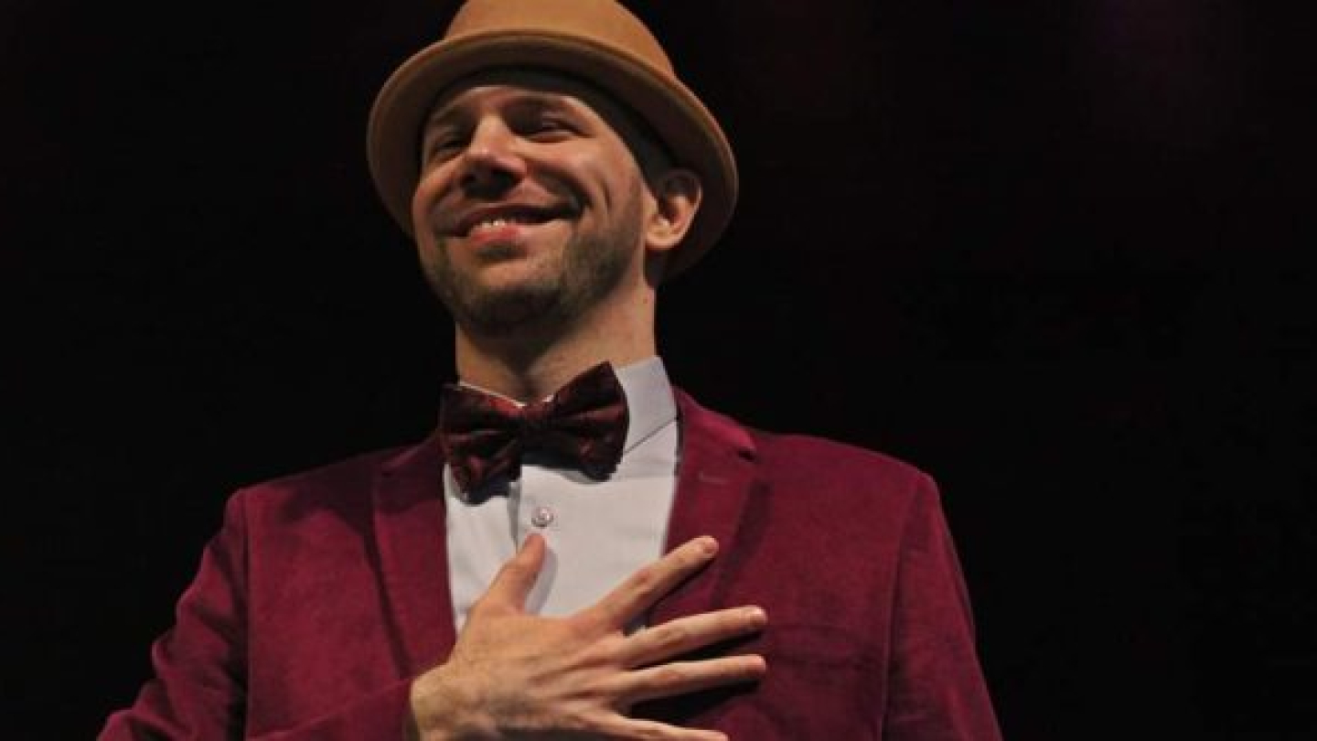 Um homem sorridente usando chapéu, gravata borboleta e paletó vinho posa com a mão no peito sobre um fundo escuro